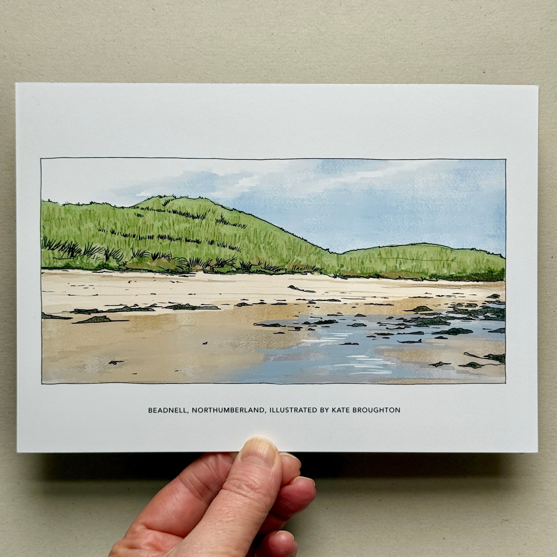 Hand holding a print of a coastal landscape with green hills, sand dunes and a beach. Labelled Beadnell, Northumberland