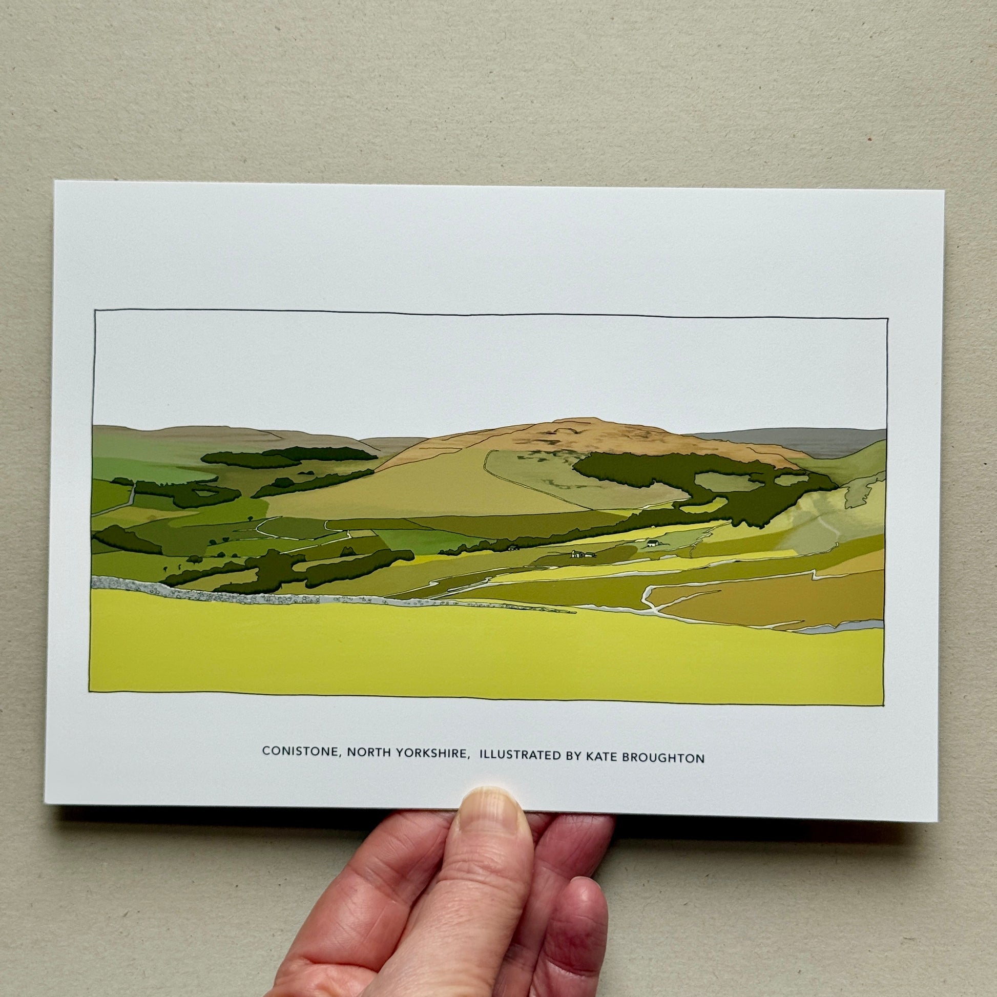 Hand holding a landscape illustration Art print with green fields and a hill on a beige background. Labelled Conistone, North Yorkshire