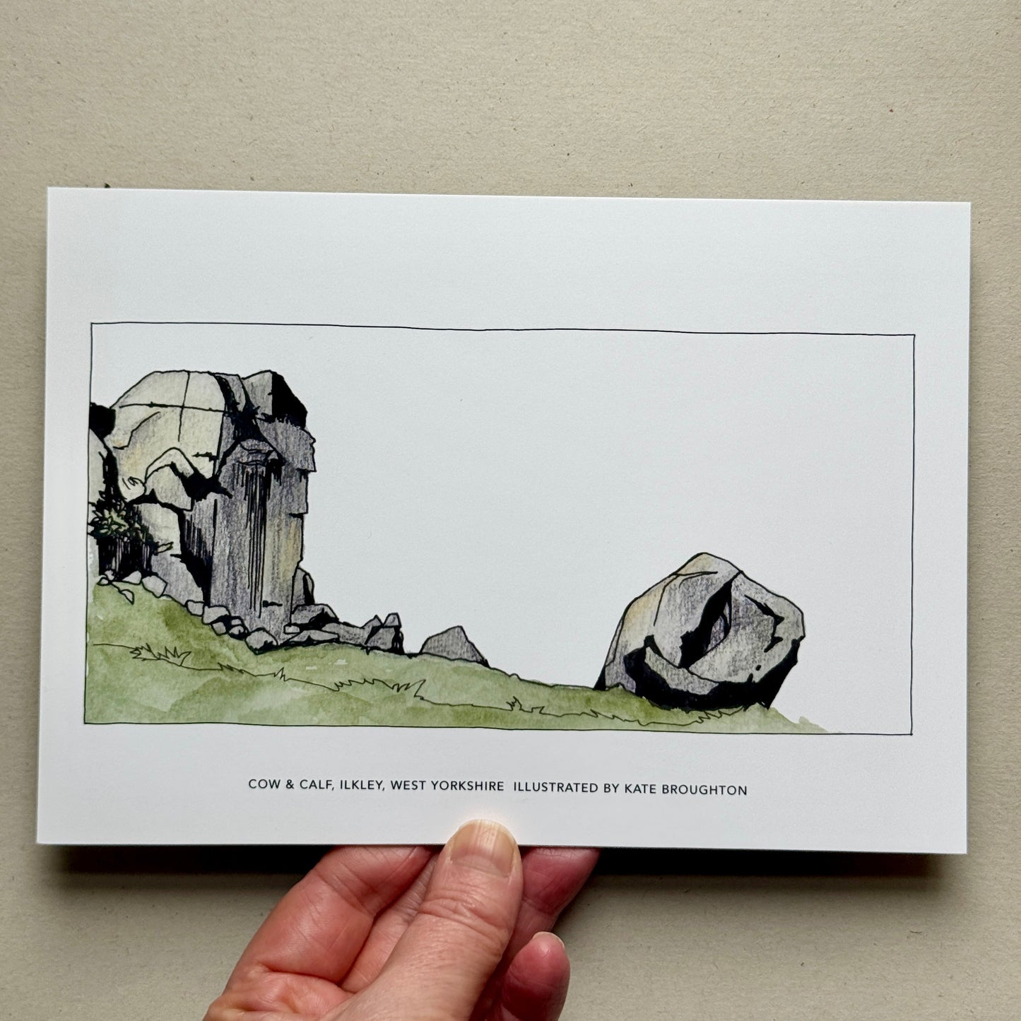 Hand holding an Art Print: Illustration of a rock formation on a white background, labelled: Cow & Calf, Ilkley, West Yorkshire.