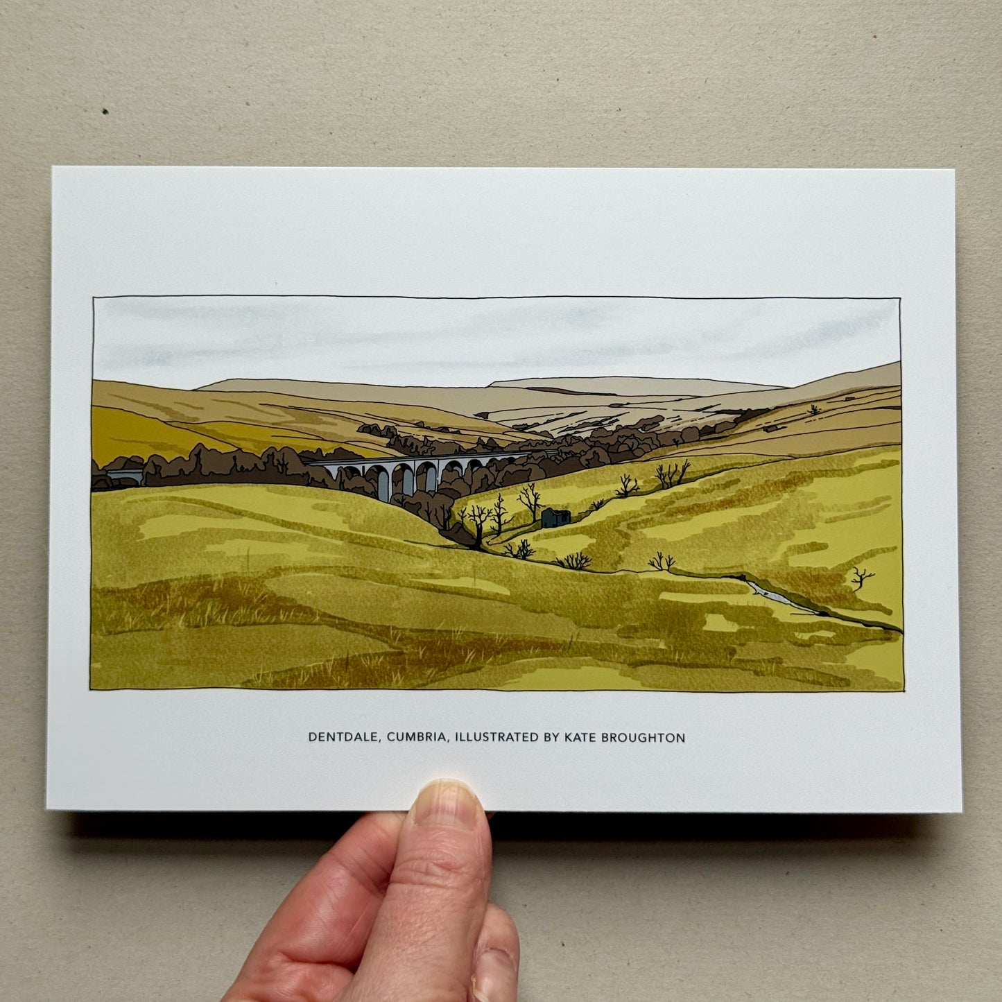 Art print of a landscape with a viaduct held by a hand on a beige background, Dental, Cumbria, by Kate Broughton