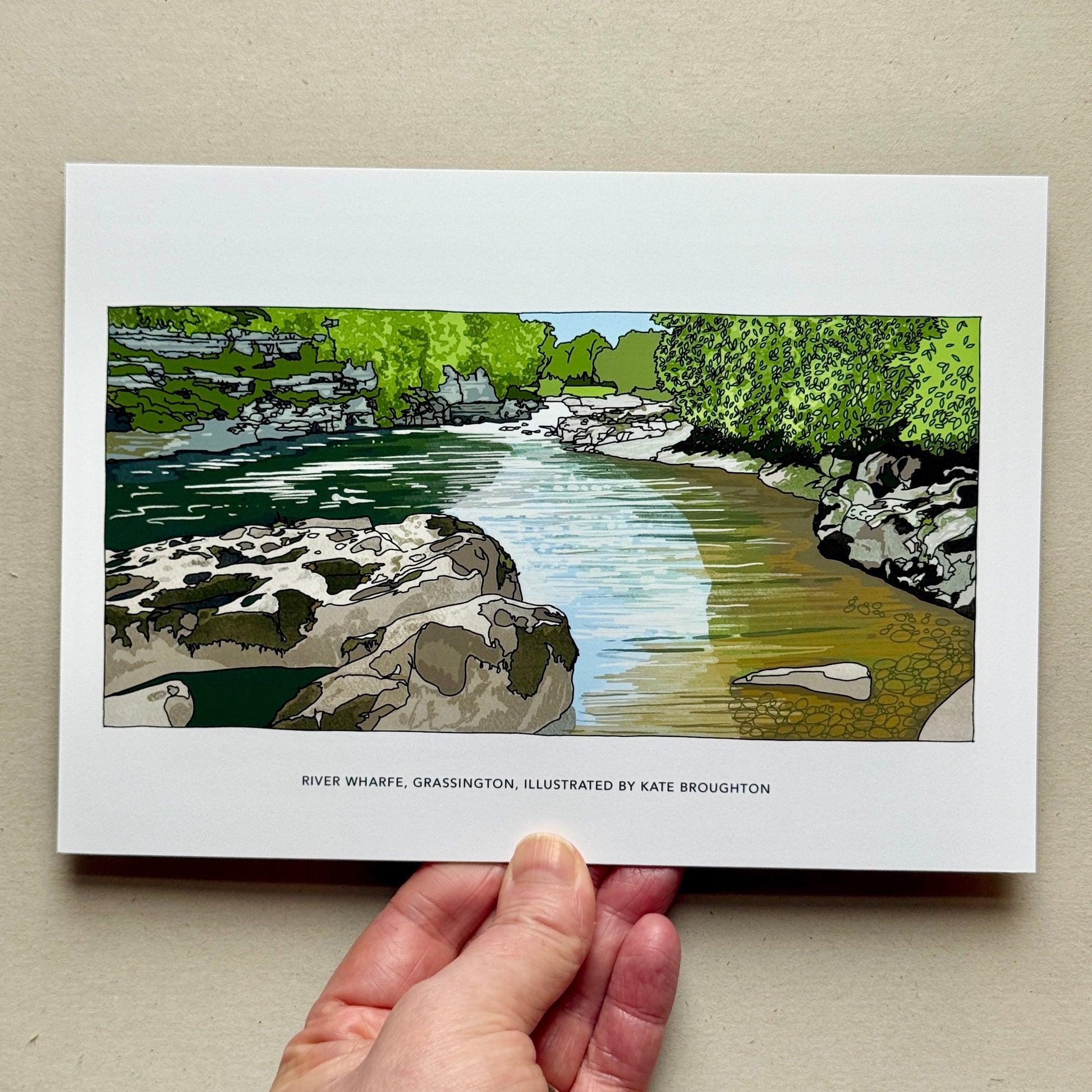 Hand holding a print of a river scene illustration on a beige background