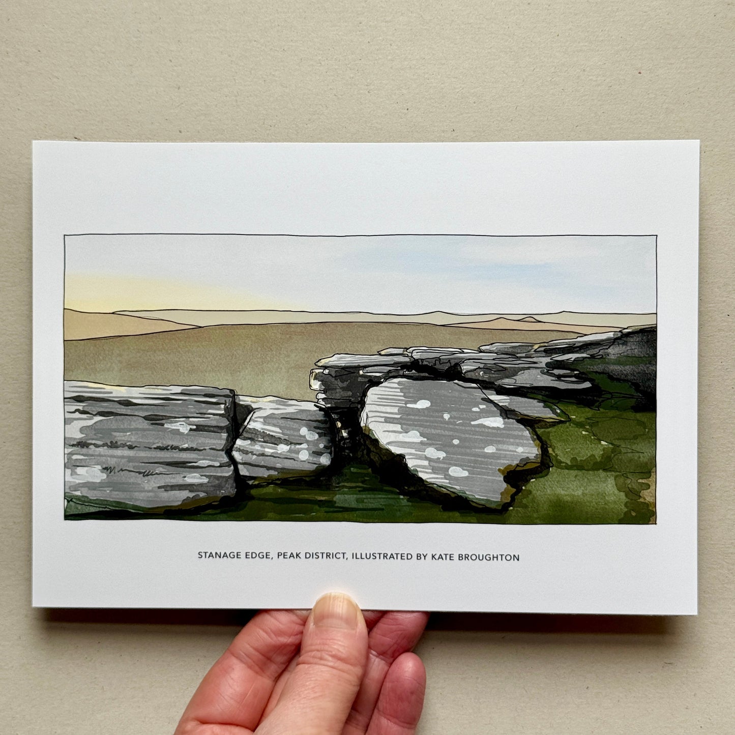 Hand holding a print of a landscape with stone structures on a beige background. Labelled: Stanage edge Peak District, illustrated by Kate Broughton