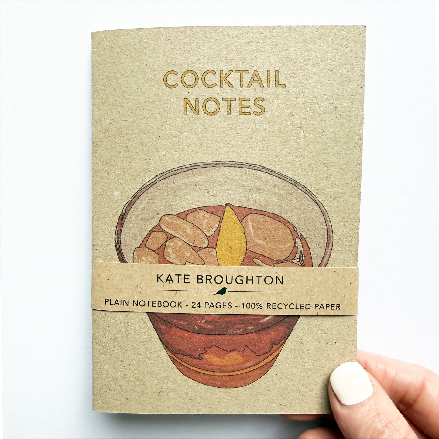 A hand holds a small kraft-paper notebook titled 'COCKTAIL NOTES' by Kate Broughton. The cover features an illustration of a cocktail glass with ice and a lemon slice, with text reading 'PLAIN NOTEBOOK - 24 PAGES - 100% RECYCLED PAPER'.
