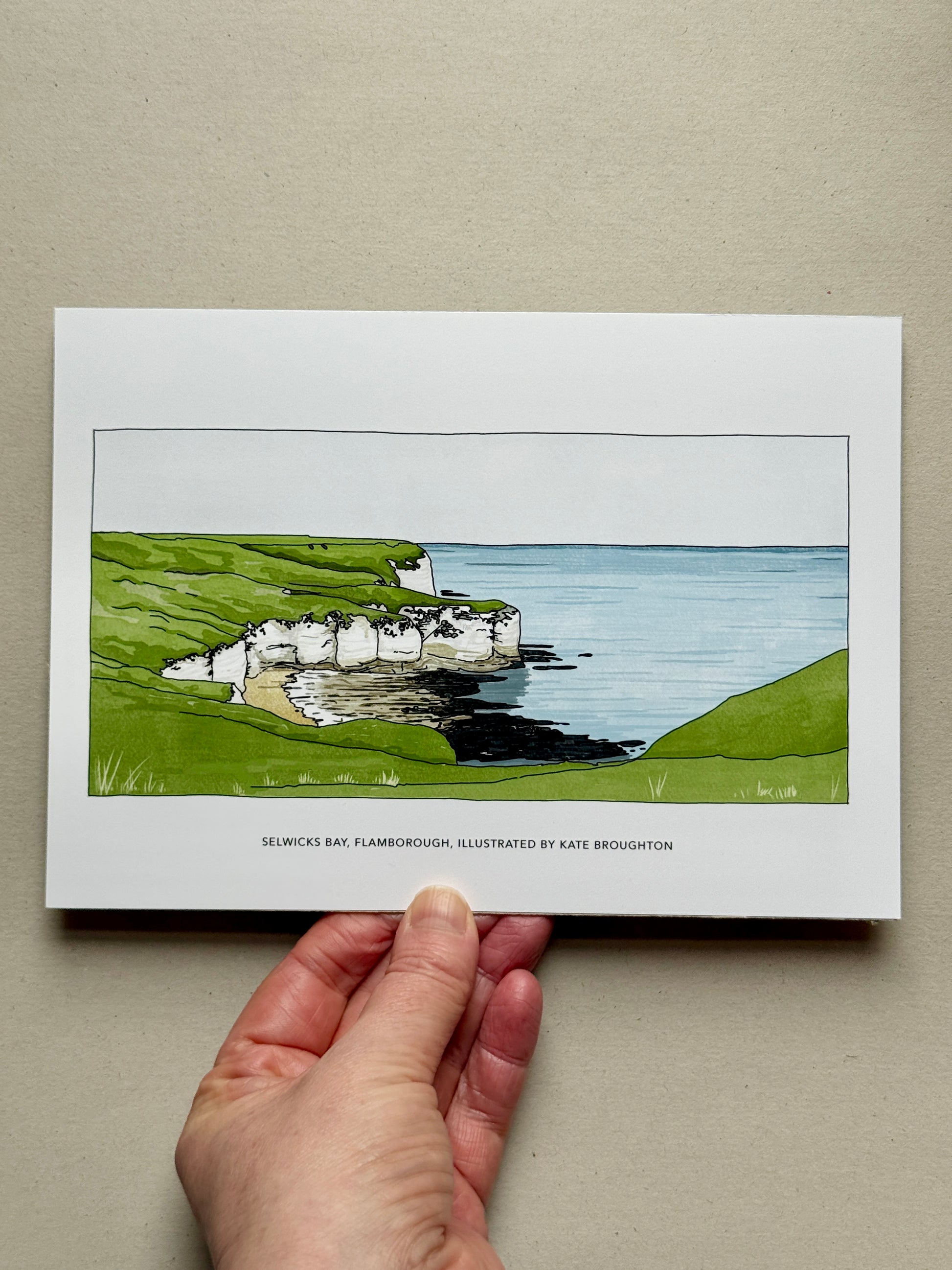 Hand holding a print of a Yorkshire coastal scene with cliffs and sea. Selwicks Bay, Flamborough by Kate Broughton